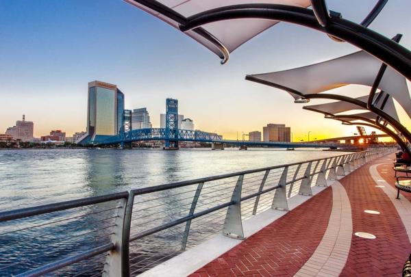 Photo of Jacksonville's 南岸河边漫步 at sunset. Red brick sidewalk alongside the river with steel guardrails. Canopies overhang the sidewalks. Jacksonville skyline in background.